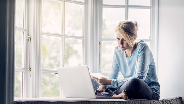 Kvinne sitter ved vindu foran sin laptop.