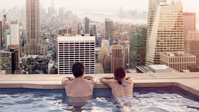 Et par som slapper av i utendørs svømmebasseng med utsikt over en storby. 